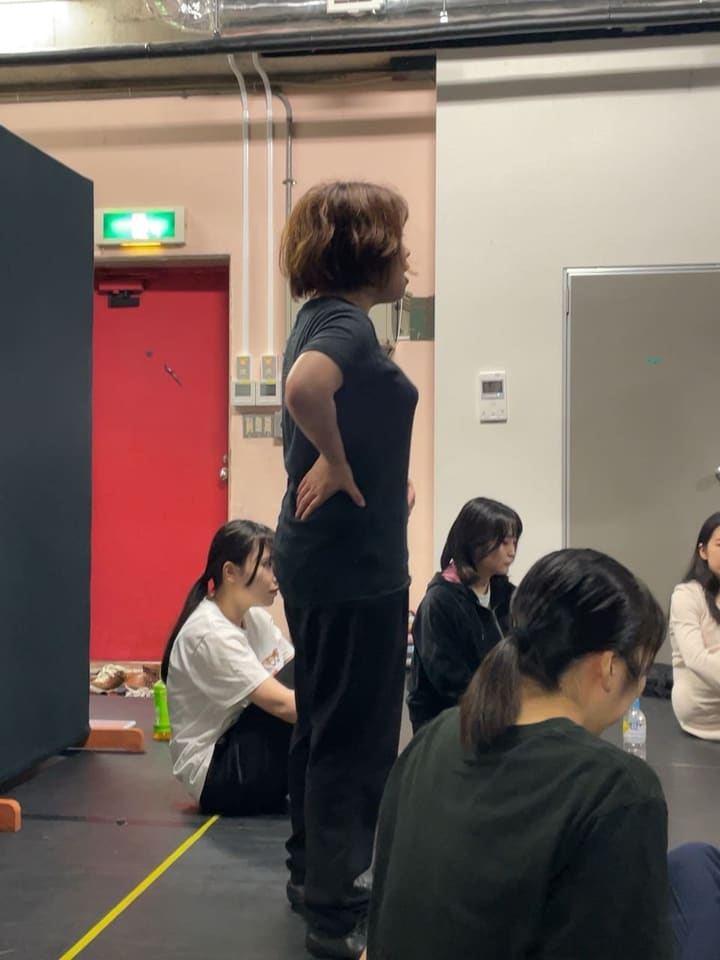腰に手を当てて立ち、座って見守る仲間の前で歌う団員|劇団カスタリア 歌稽古