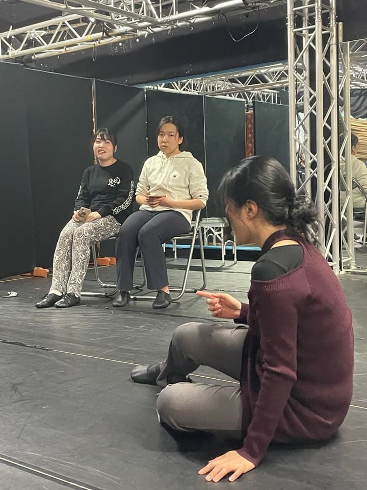 床に座って指を差し演じる出演者と、椅子で見守る団員たち|劇団カスタリア 立ち稽古