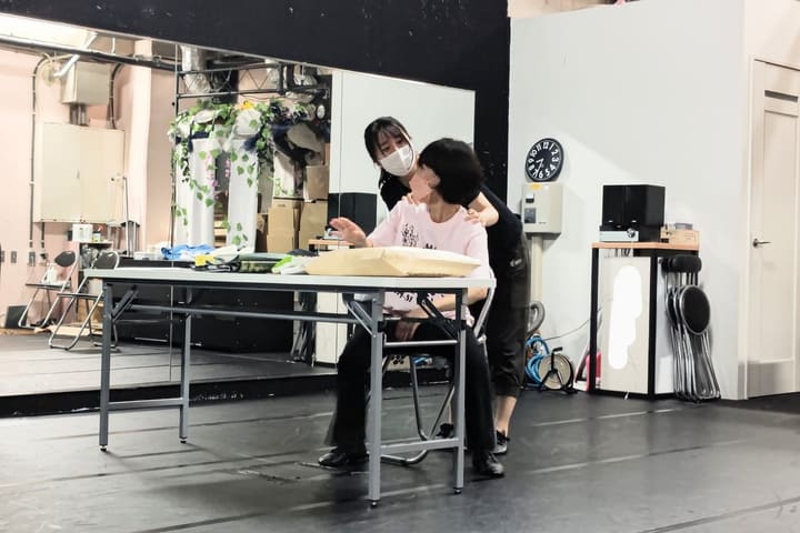 机に着いた相手の肩に手を置いて話しかける出演者|劇団カスタリア 二人芝居の稽古