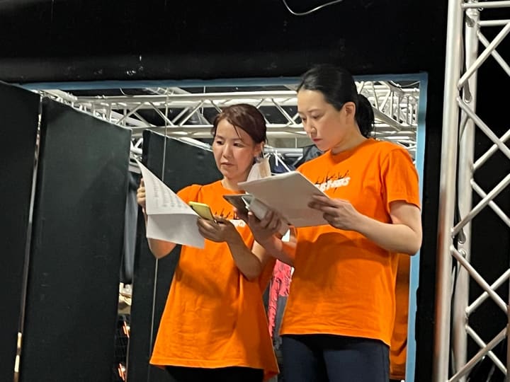 そろいのオレンジのTシャツで新曲の譜面を読み込む団員2人|劇団カスタリア「TRIP」歌稽古