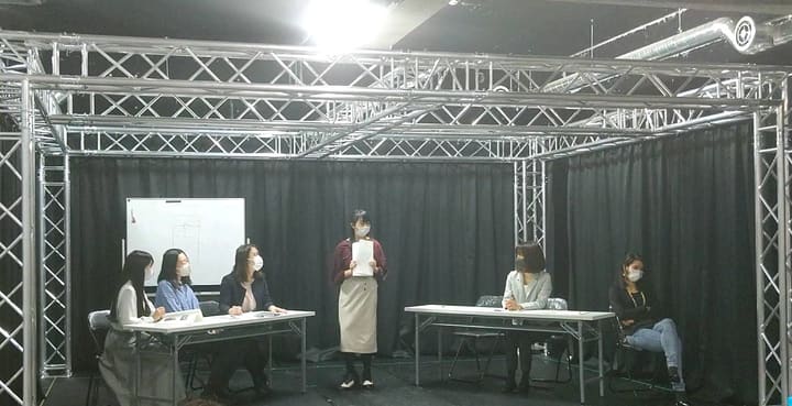 長机に着いた出演者の前で、書類を手に立って話す出演者｜劇団カスタリア「ジャッジメント」芝居稽古