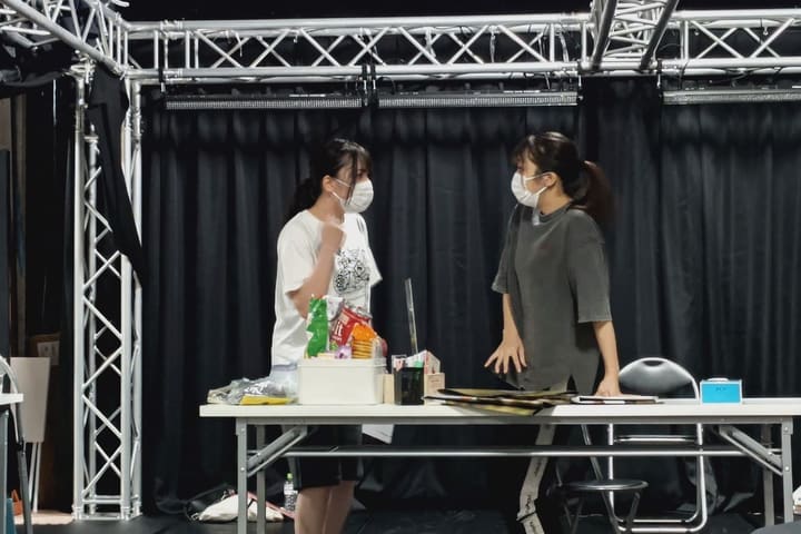 菓子や紙類を並べた長机を挟んで向き合う団員2人｜劇団カスタリア 芝居の稽古