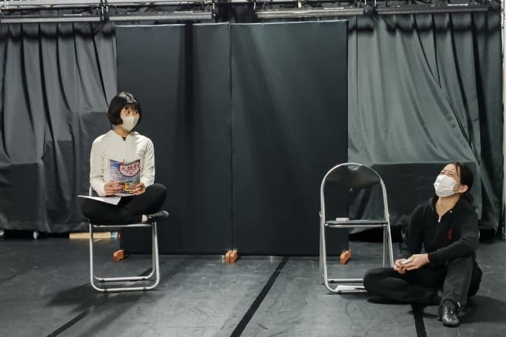 椅子の上であぐらをかき冊子を持つ団員と、床に座る相手役｜劇団カスタリア 演技稽古