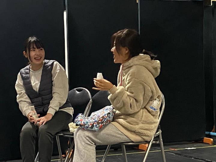 パイプ椅子に並んで座り、笑顔で言葉を交わす二人芝居の場面|劇団カスタリア 芝居稽古