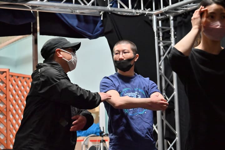 腕を組んで立つ出演者に、隣の人が身振りで動きを説明する｜劇団カスタリア「オーブ」稽古