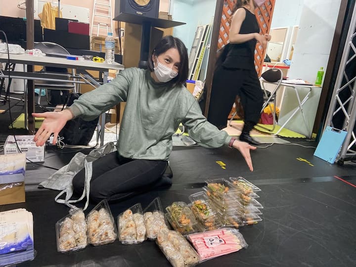 床に並んだおにぎりと惣菜の差し入れを両手で示す団員｜劇団カスタリア 稽古オフショット