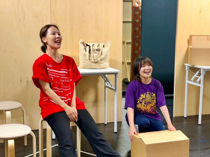 段ボール箱のそばに座り、顔を見合わせて笑う2人の出演者｜劇団カスタリア 稽古の合間