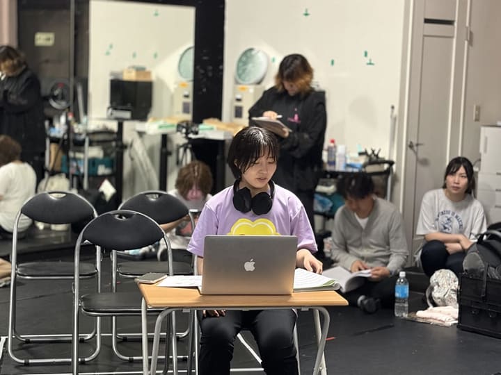 稽古場の机でノートパソコンに向かい制作作業を進める団員と、床で台本を読み込むメンバー|劇団カスタリア「金曜日の食卓」稽古