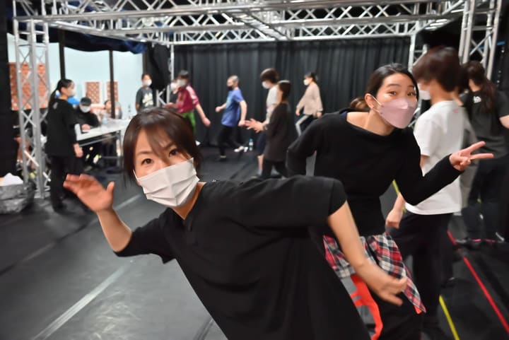 大勢が踊る稽古場の手前で、カメラに向かってポーズをとる2人｜劇団カスタリア 稽古オフショット