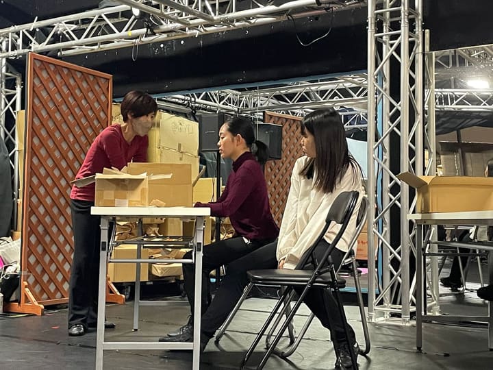 段ボール箱が積まれた場面で、机の奥に立つ団員と椅子に座る2人が向き合う|劇団カスタリア 通し稽古
