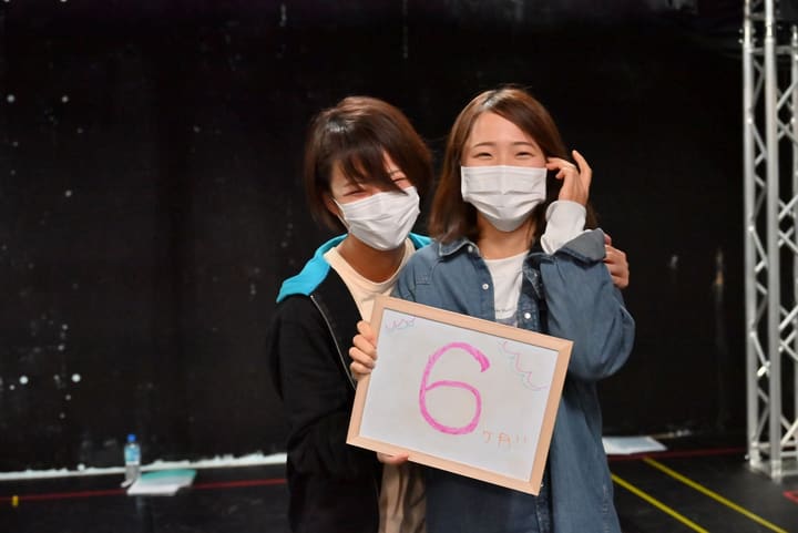 「6ヶ月!!」と書いたホワイトボードを掲げて笑う団員2人|劇団カスタリア 稽古オフショット