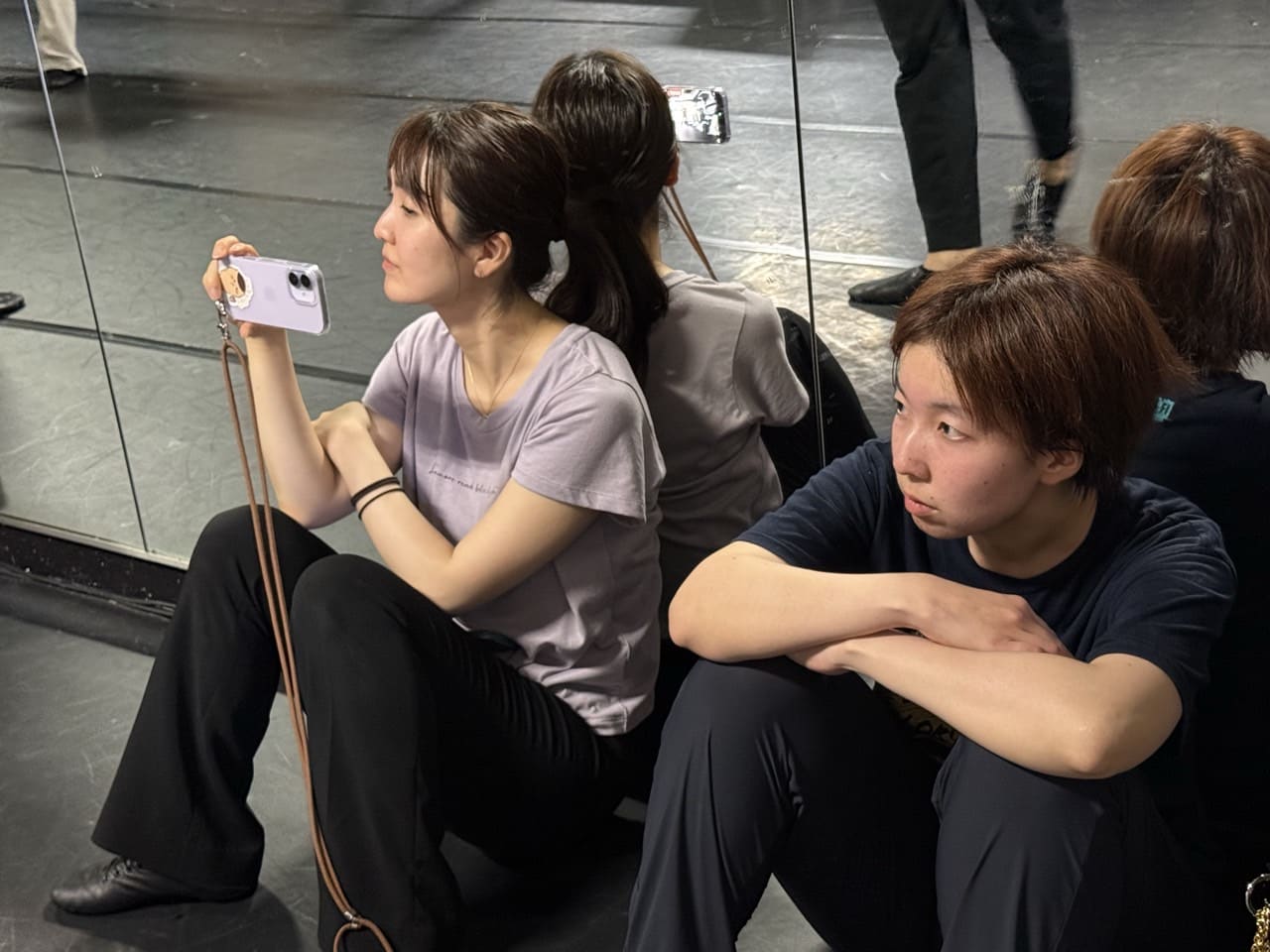 鏡張りの稽古場の床に座り、スマートフォンで振付を撮影しながら見守る団員たち｜劇団カスタリア「金曜日の食卓」稽古